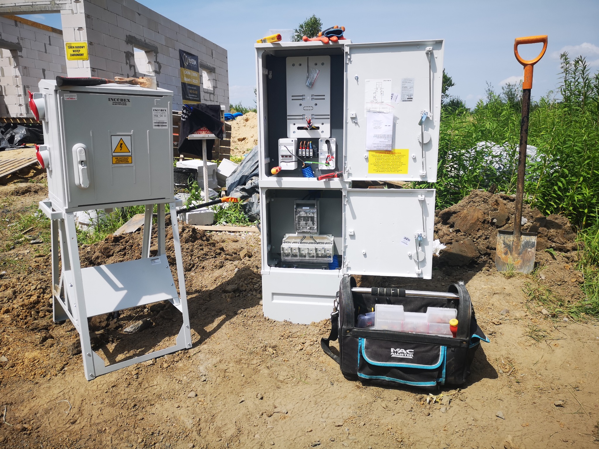 Dwa rozdzielcze panele elektryczne IncoBex na placu budowy, otwarte drzwi odsłaniają wnętrze jednego z paneli, obok stoi torba narzędziowa Mac Allister i wbita w ziemię łopata