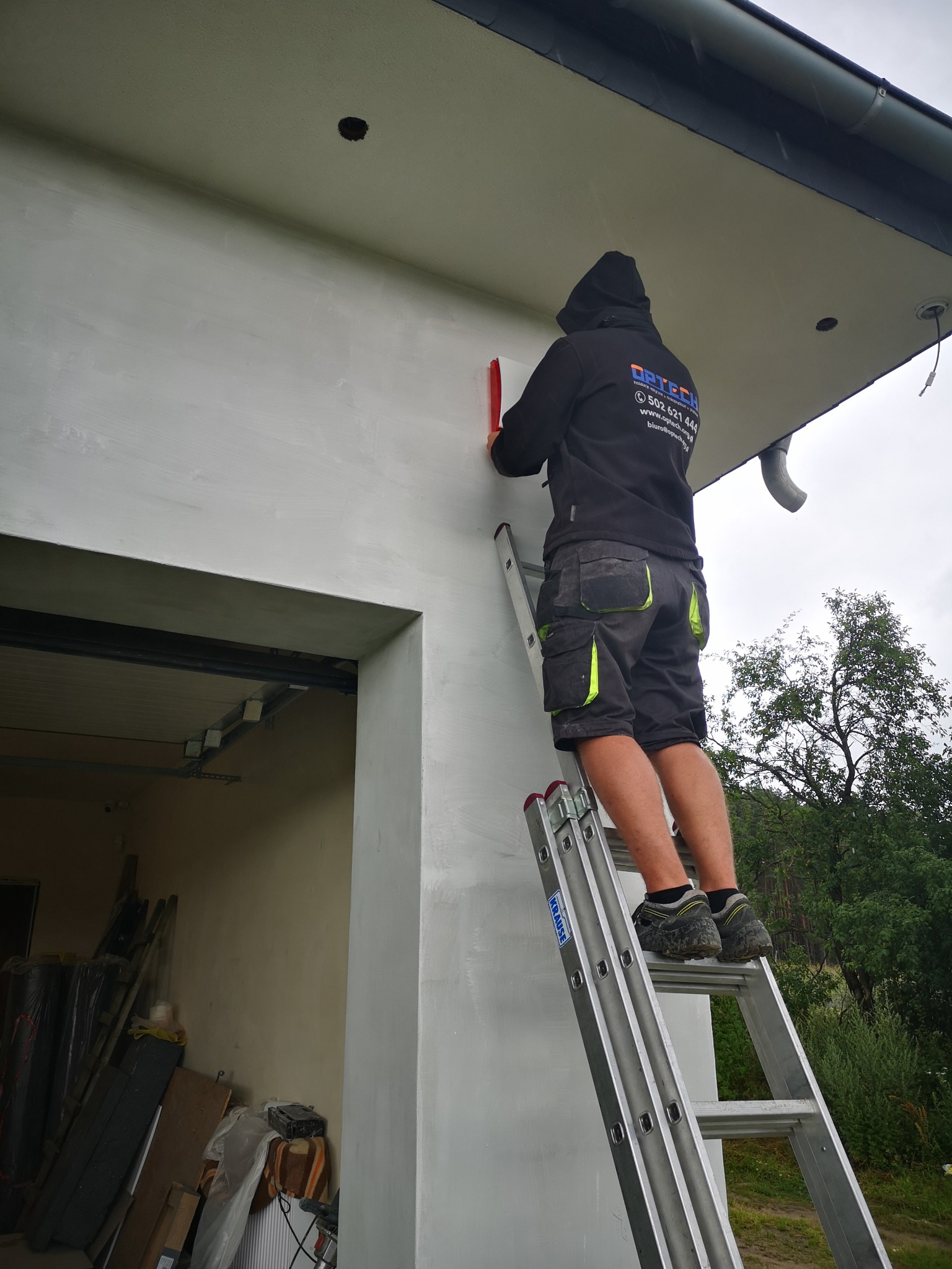 Mężczyzna w bluzie z kapturem na drabinie, przykłada poziomicę do świeżo otynkowanej ściany budynku z widocznymi elementami instalacji elektrycznej i rynną.