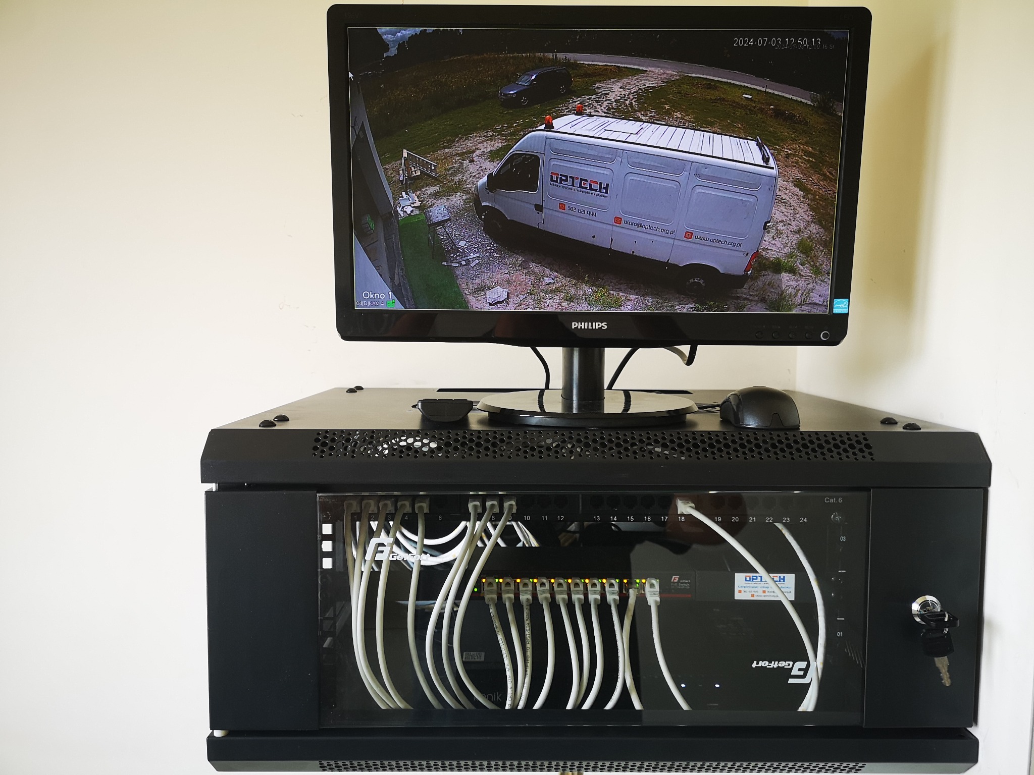Monitor wyświetla obraz z kamery monitoringu przedstawiający biały samochód dostawczy firmy Optech na żwirowym podjeździe. Poniżej szafa rack z patch panelem i podłączonymi kablami Ethernet kat. 6.