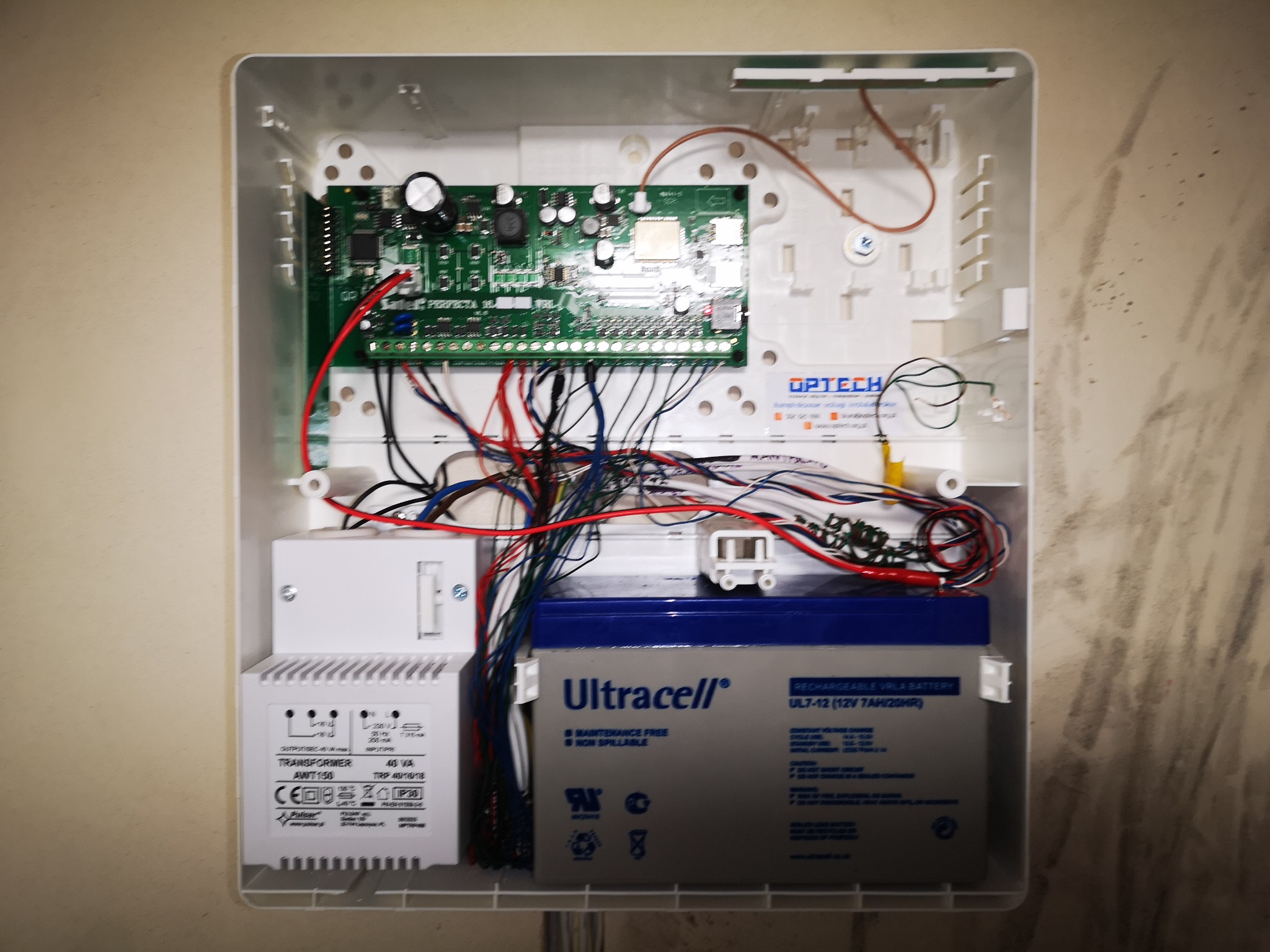 Otwarta obudowa centrali alarmowej z widoczną płytą główną, transformatorem, akumulatorem Ultracell i plątaniną przewodów wewnątrz.