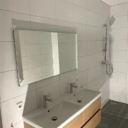 Nowoczesna łazienka z podwójną umywalką w drewnianej szafce, lustrem i prysznicem na tle białych płytek, widoczne zabezpieczenia folią na elementach armatury.