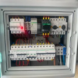 Otwarta skrzynka rozdzielcza z widocznymi bezpiecznikami, stycznikami i przewodami w różnych kolorach, z detalami marki Schneider i Eaton, oraz czerwoną diodą sygnalizacyjną na jednym z elementów.