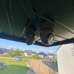 Dwie kamery obrotowe marki PIXIR zamontowane pod zadaszeniem, widok na posesję z domami i zielenią w tle.