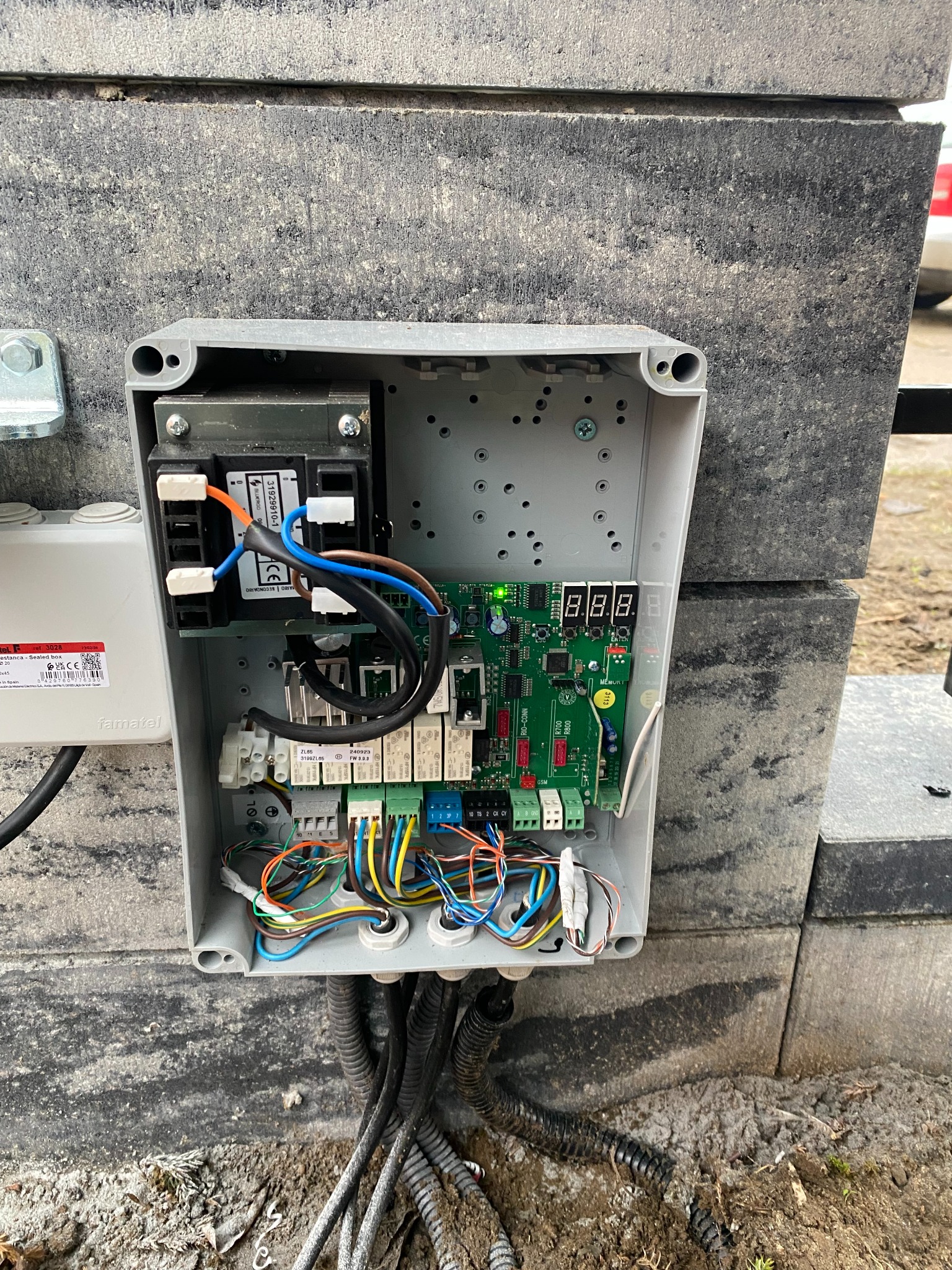 Otwarta skrzynka sterownicza automatu bramy z widocznymi przewodami, płytką drukowaną i wyświetlaczem LED, zamontowana na szarym, betonowym słupku ogrodzeniowym.