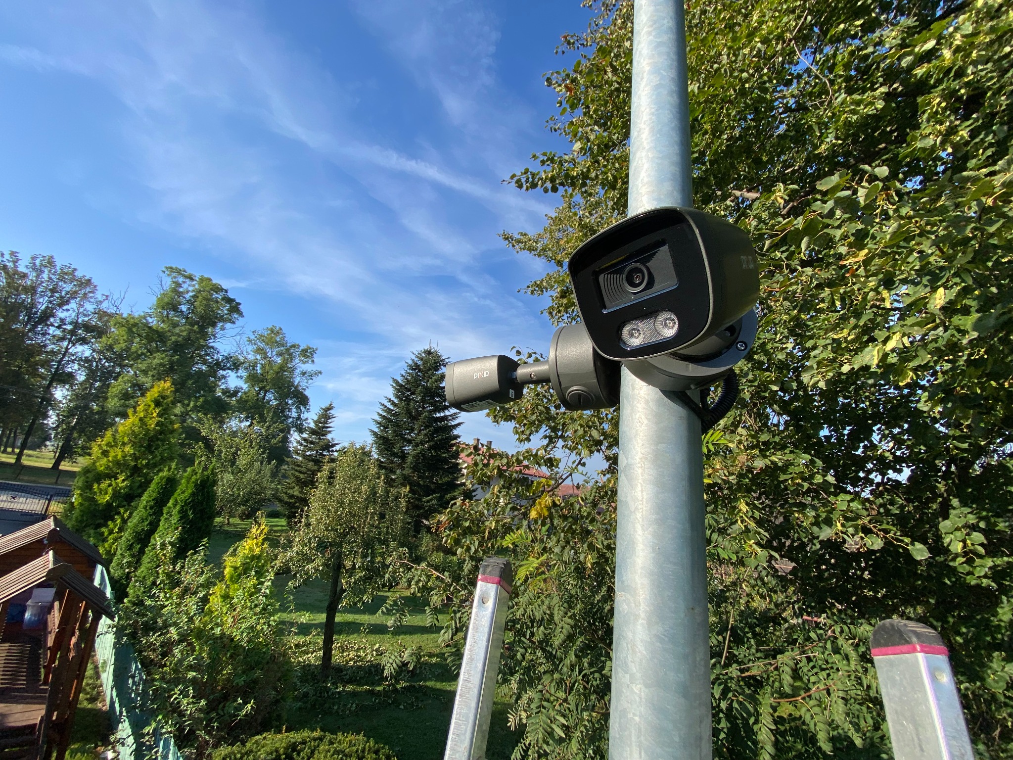 Kamera monitoringu zewnętrznego zamontowana na metalowym słupie, widok z dołu na tle błękitnego nieba i zieleni ogrodu, drabina oparta o słup.