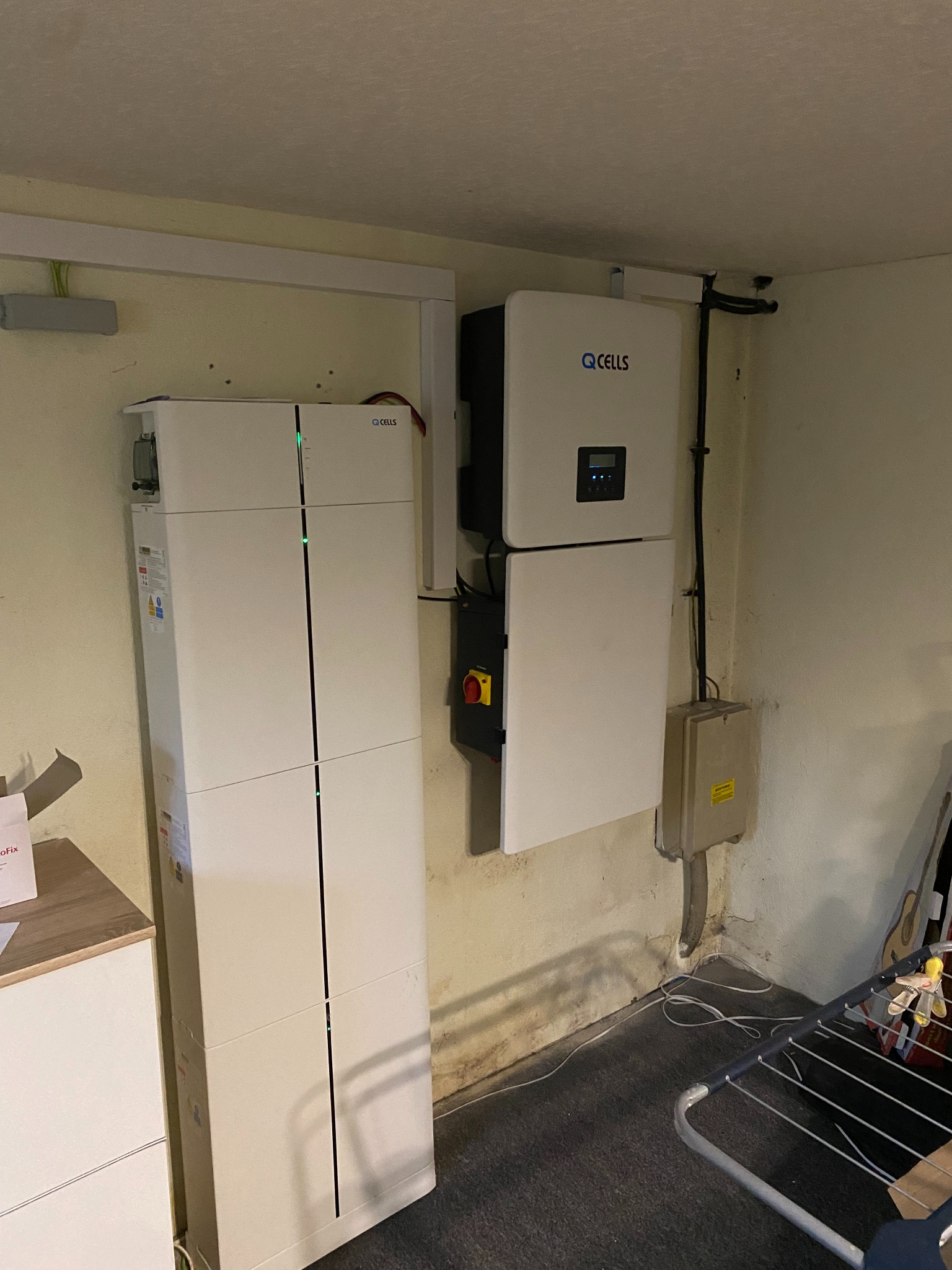 Biała bateria magazynująca energię Q CELLS zamontowana na ścianie w pomieszczeniu, obok widoczny inwerter tej samej firmy, instalacja elektryczna z przewodami i skrzynką przyłączeniową.