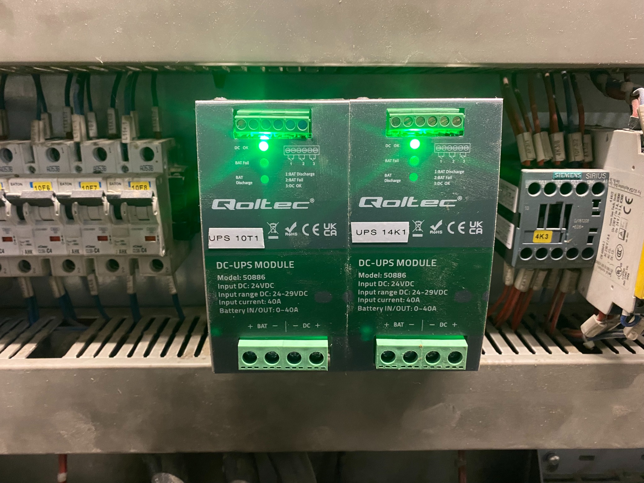 Dwa moduły DC-UPS firmy Qoltec (modele 50886, UPS 10T1 i UPS 14K1) zamontowane w szafie elektrycznej, z widocznymi diodami LED wskazującymi status pracy i podłączeniami kablowymi, obok wyłączników...