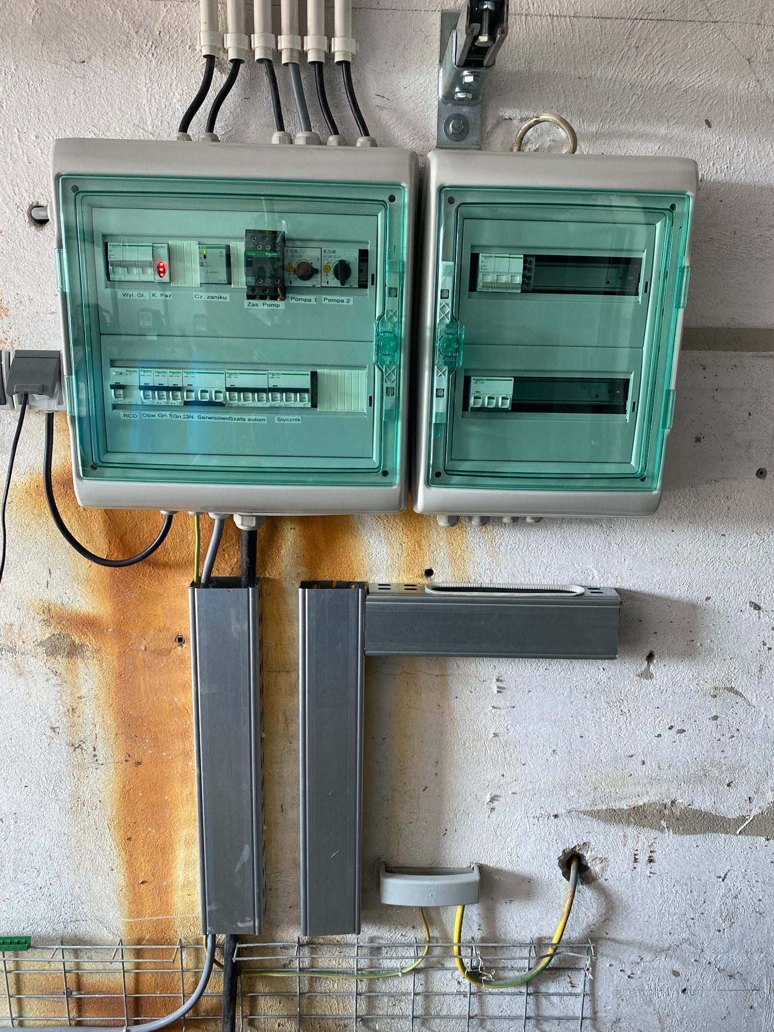 Dwa panele elektryczne z zielonymi obudowami zamontowane na szarej, częściowo pomalowanej ścianie z widocznymi przewodami i elementami montażowymi.
