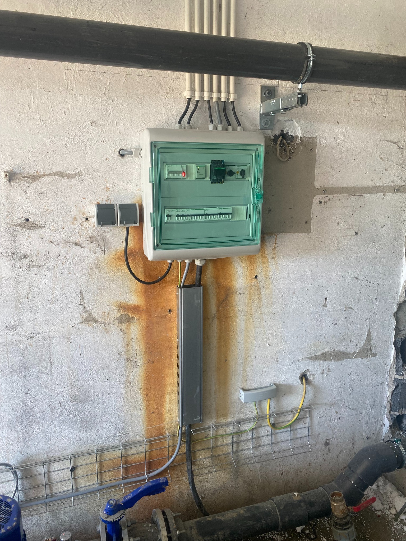 Zamontowana skrzynka elektryczna z zielonymi elementami, widoczna na tle surowej ściany z zaciekami, z przewodami wchodzącymi do skrzynki od góry i dołu, oraz fragment instalacji hydraulicznej...