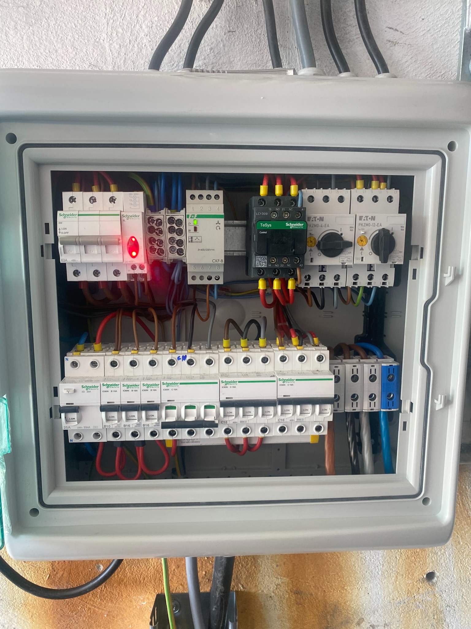 Otwarta skrzynka rozdzielcza z widocznymi bezpiecznikami, stycznikami i przewodami w różnych kolorach, z detalami marki Schneider i Eaton, oraz czerwoną diodą sygnalizacyjną na jednym z elementów.