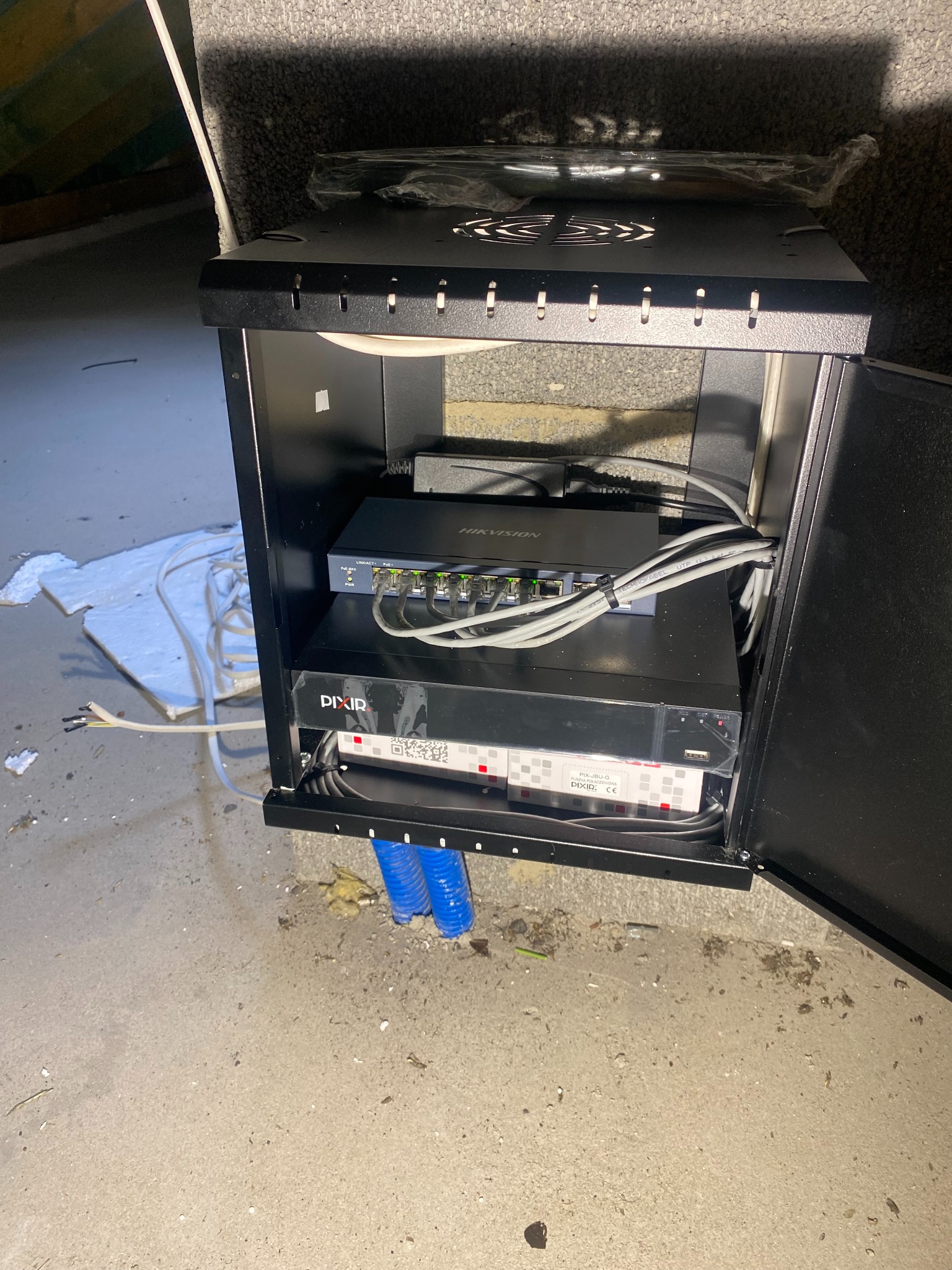 Czarna szafka rack z zamontowanym switchem Hikvision, rejestratorem Pixir i przewodami, widoczne niebieskie rury instalacyjne w podłodze.