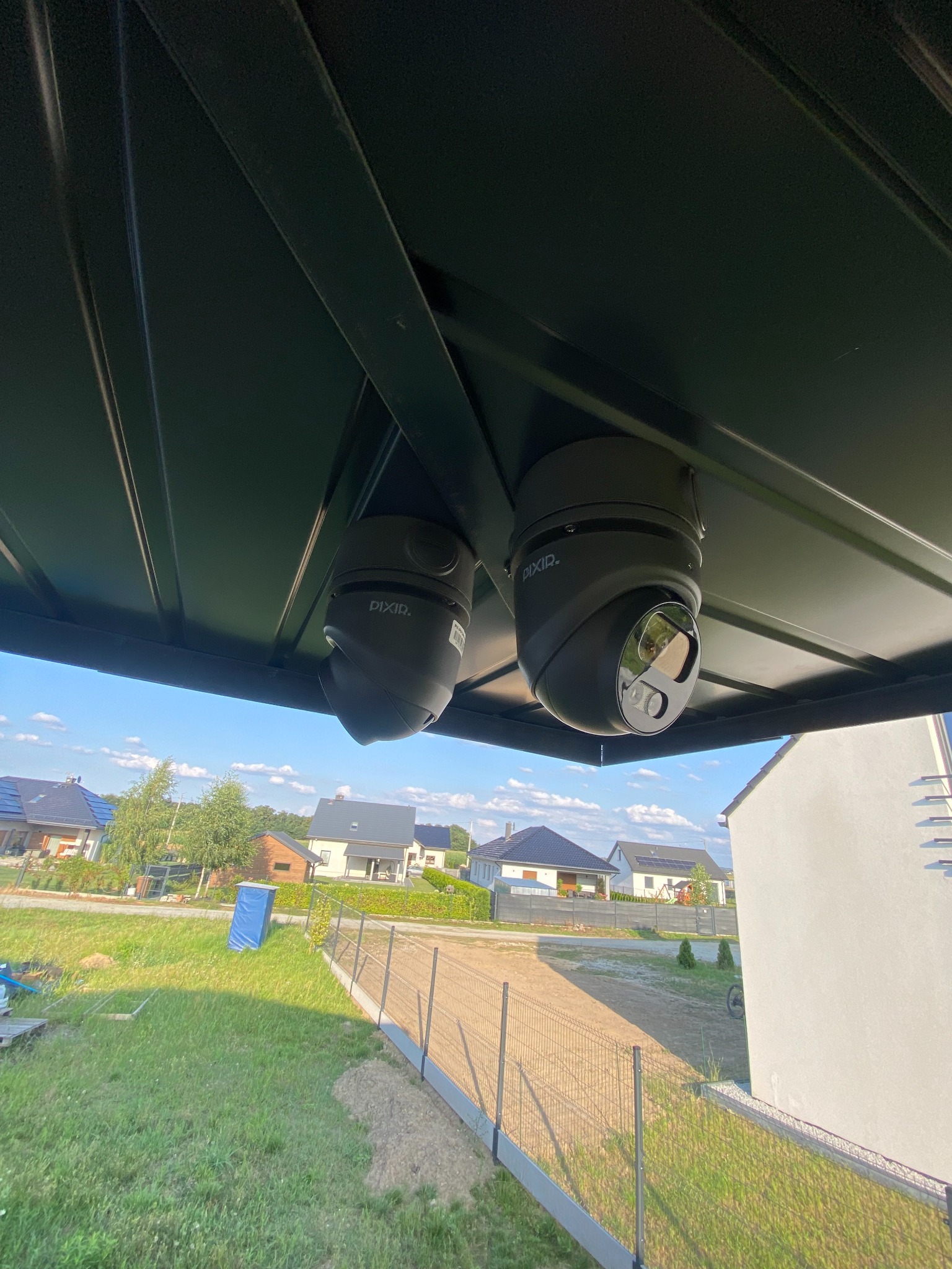 Dwie kamery obrotowe marki PIXIR zamontowane pod zadaszeniem, widok na posesję z domami i zielenią w tle.
