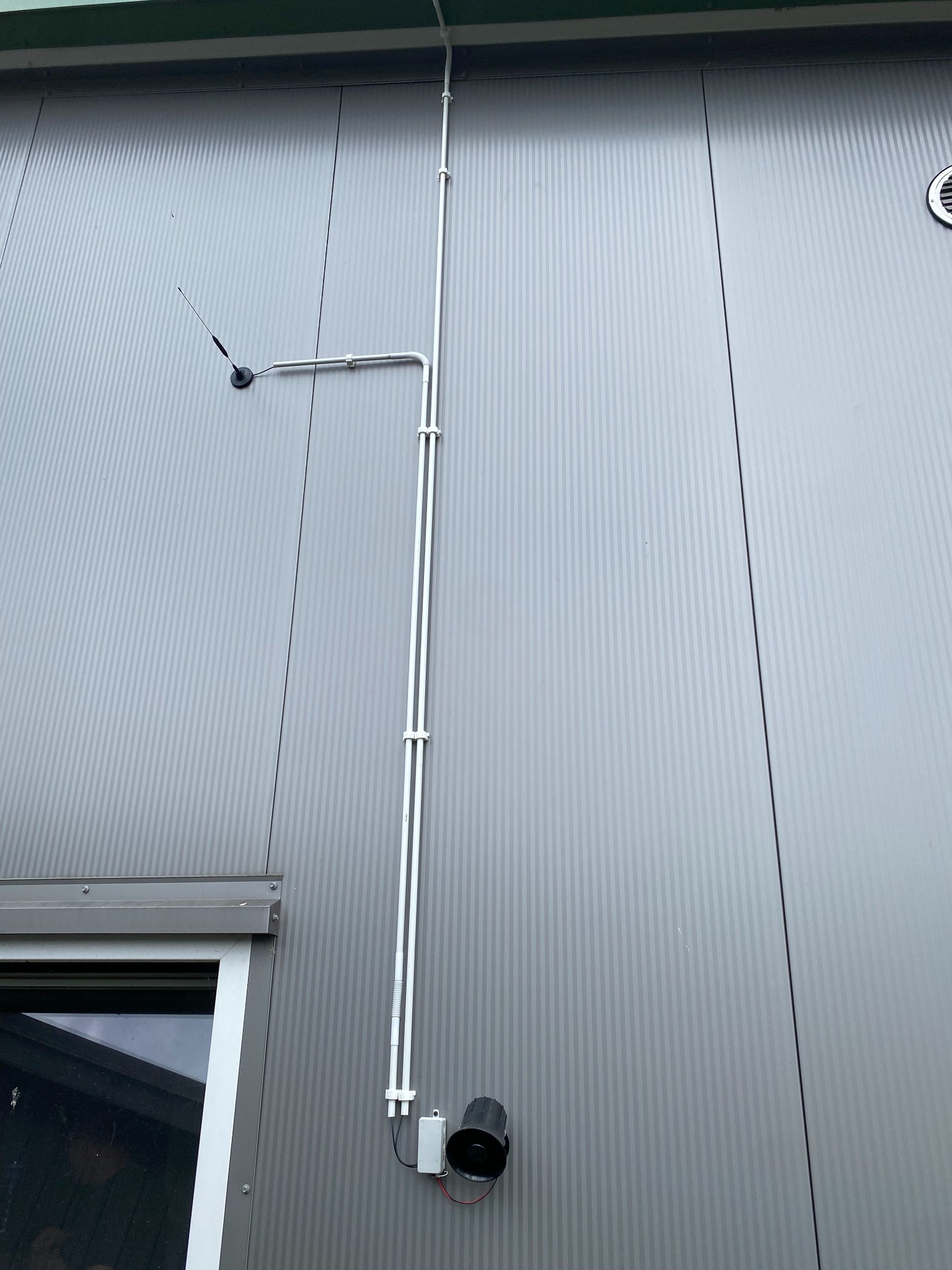 Zewnętrzny montaż anteny i syreny alarmowej na szarej elewacji budynku w Wieluniu, z widocznymi przewodami w białych rurkach PCV.