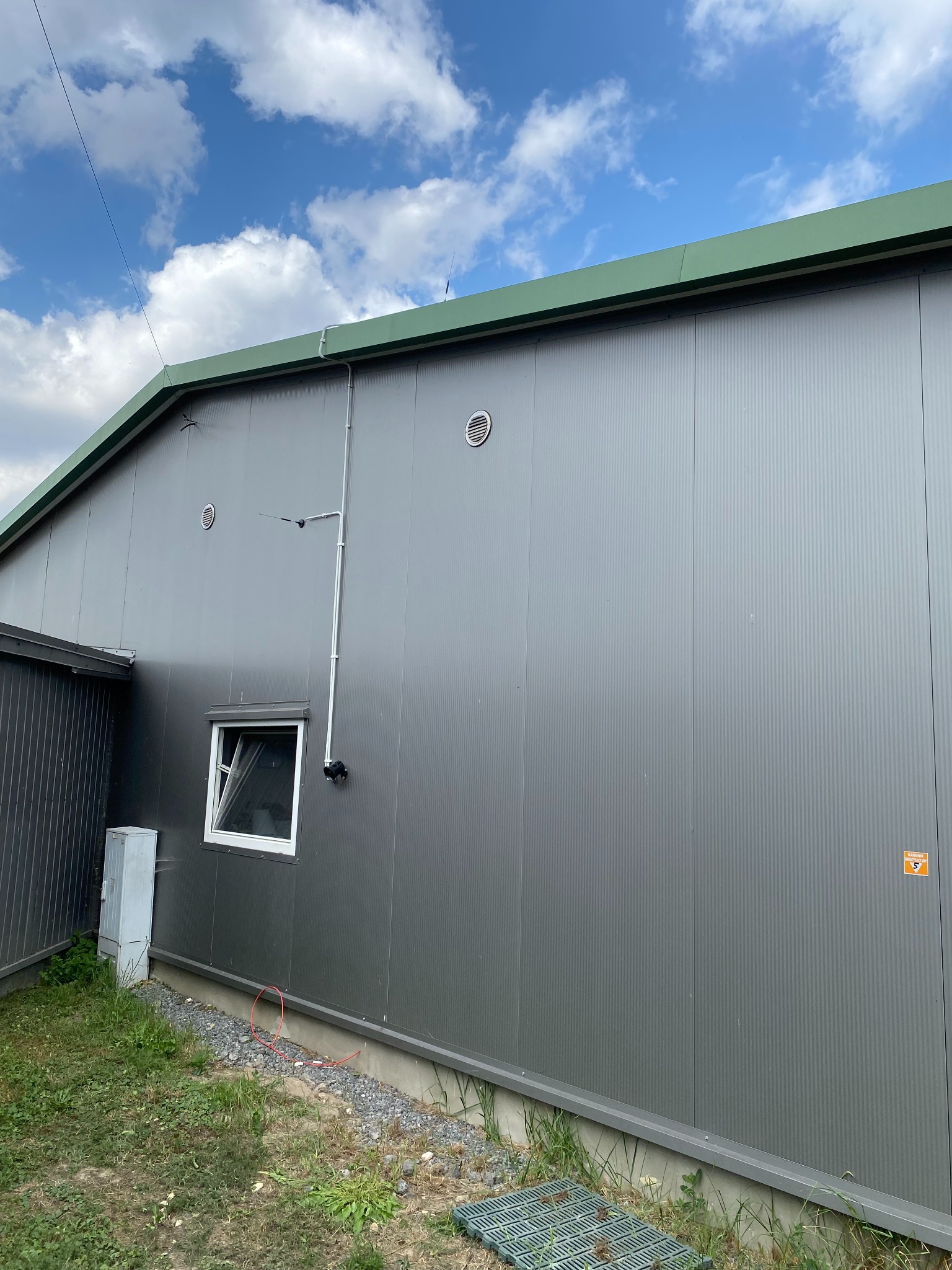 Szara hala przemysłowa z oknem, wentylacją, anteną i rurami na elewacji, widoczna skrzynka elektryczna przy ziemi i fragment czerwonego kabla.