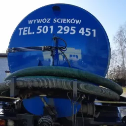 Niebieski zbiornik asenizacyjny z numerem telefonu do wywozu nieczystości, widok z tyłu, na tle drzew i budynku.