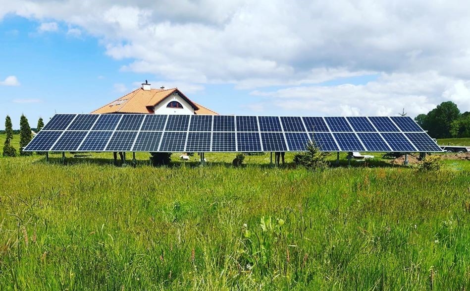 Instalacja paneli słonecznych na konstrukcji wsporczej, z domem w tle, otoczona wysoką trawą na słonecznym terenie.