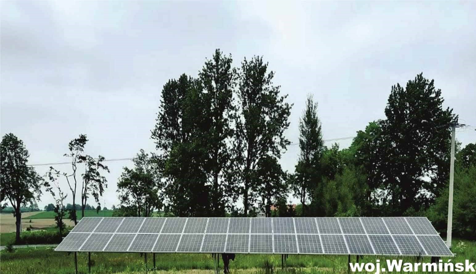 Rząd paneli fotowoltaicznych na tle zieleni i pochmurnego nieba w województwie warmińsko-mazurskim.