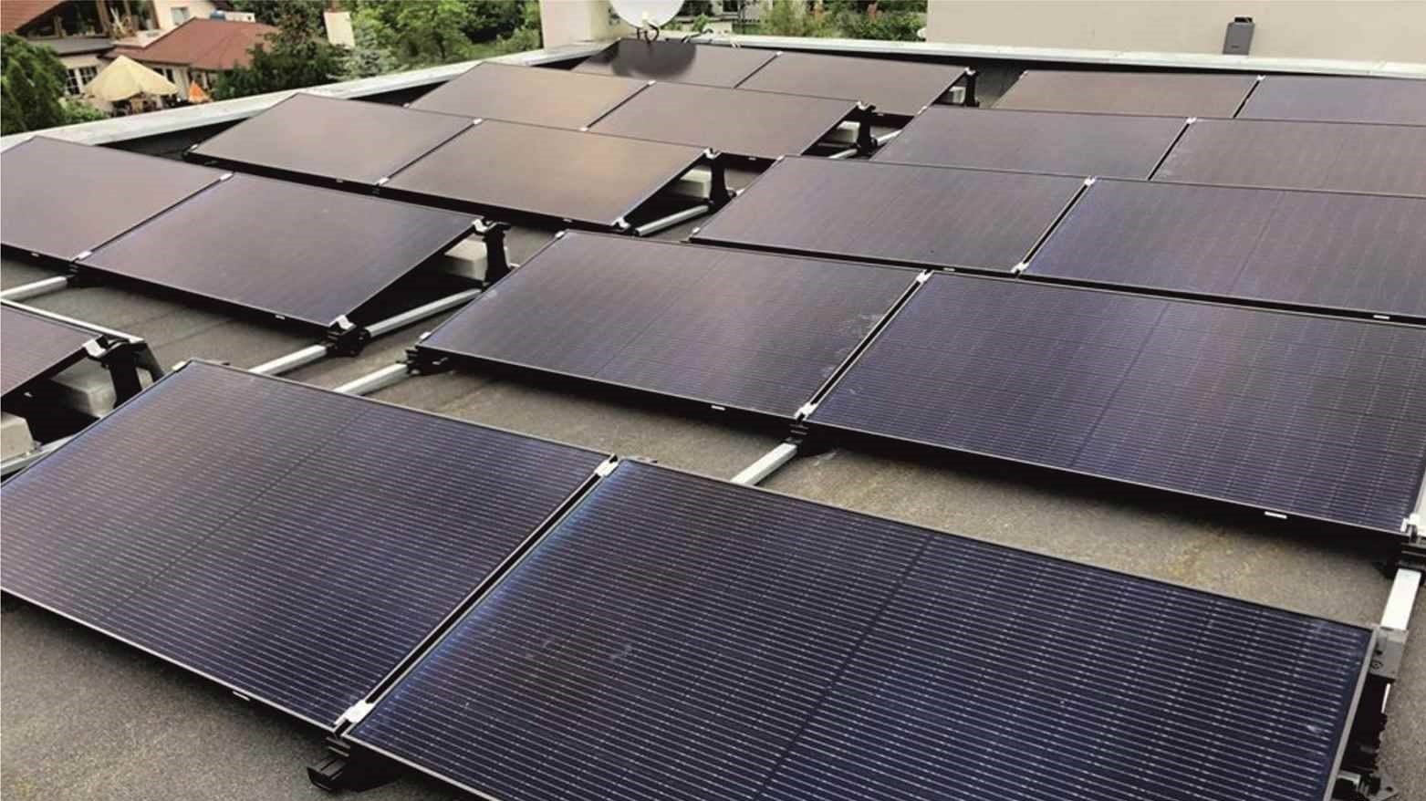 Rząd paneli słonecznych na płaskim dachu budynku mieszkalnego, widoczne białe mocowania i antena satelitarna w tle.