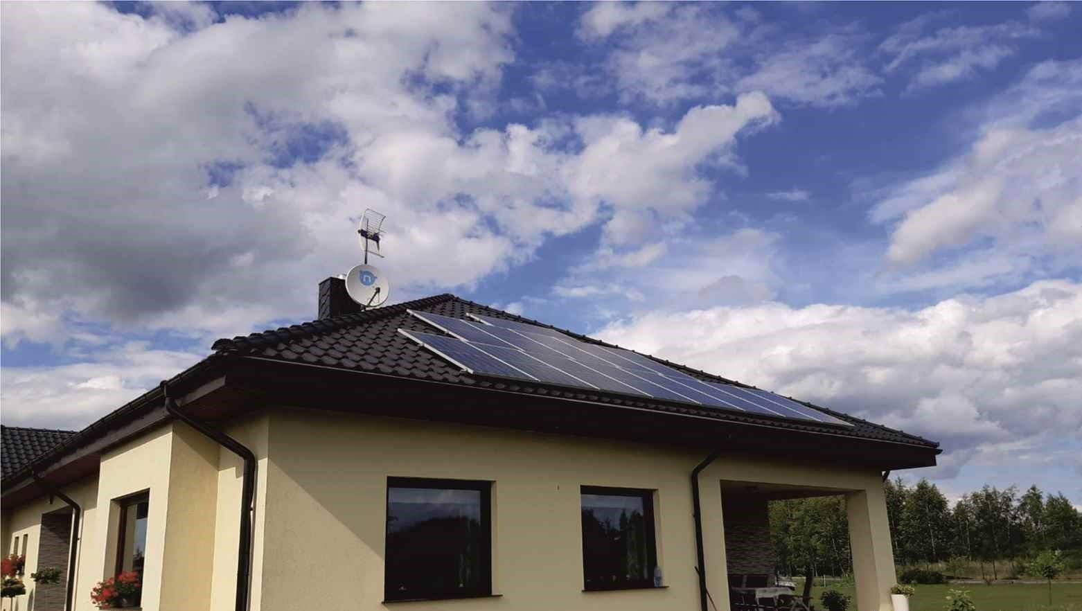 Panele słoneczne na dachu domu jednorodzinnego z anteną satelitarną i telewizyjną, błękitne niebo z obłokami w tle.
