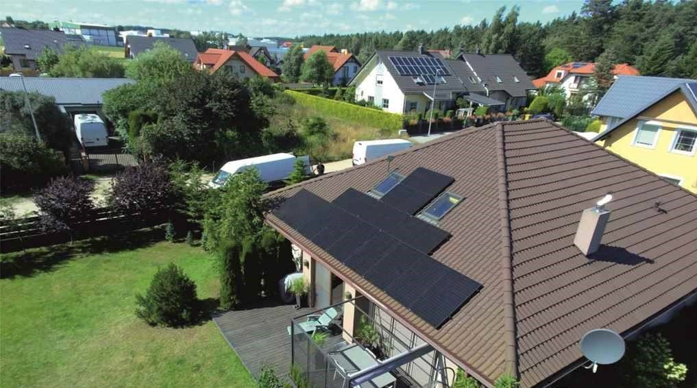 Instalacja paneli słonecznych na brązowym dachu domu jednorodzinnego z widocznymi oknami dachowymi, w tle zabudowa mieszkalna i zieleń, ujęcie z góry.