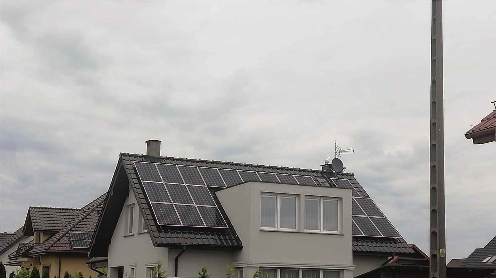 Panele fotowoltaiczne na dachu domu jednorodzinnego z anteną satelitarną i kominem, widok z poziomu ulicy w pochmurny dzień.