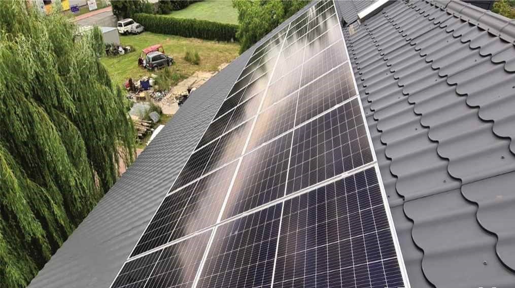 Panele słoneczne zamontowane na dachu pokrytym szarą dachówką ceramiczną, widok z góry na instalację i otoczenie z zielenią.
