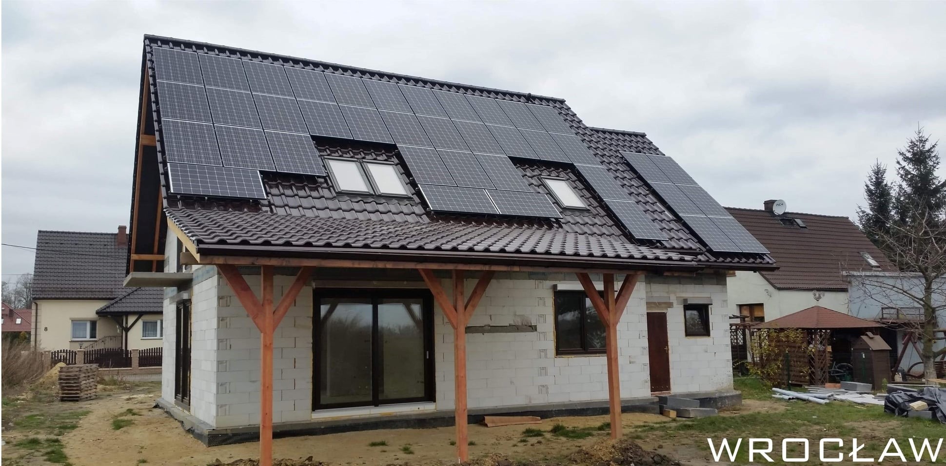 Dom jednorodzinny w trakcie budowy z zainstalowanymi panelami słonecznymi na brązowym dachu. Widoczne okna dachowe i drewniana konstrukcja werandy. Na pierwszym planie gruz budowlany. W tle...