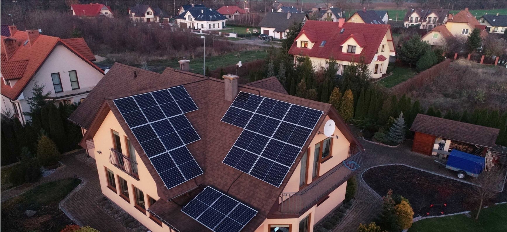 Widok z góry na dom z brązowym dachem, na którym zamontowane są panele słoneczne. W tle inne domy i drzewa.