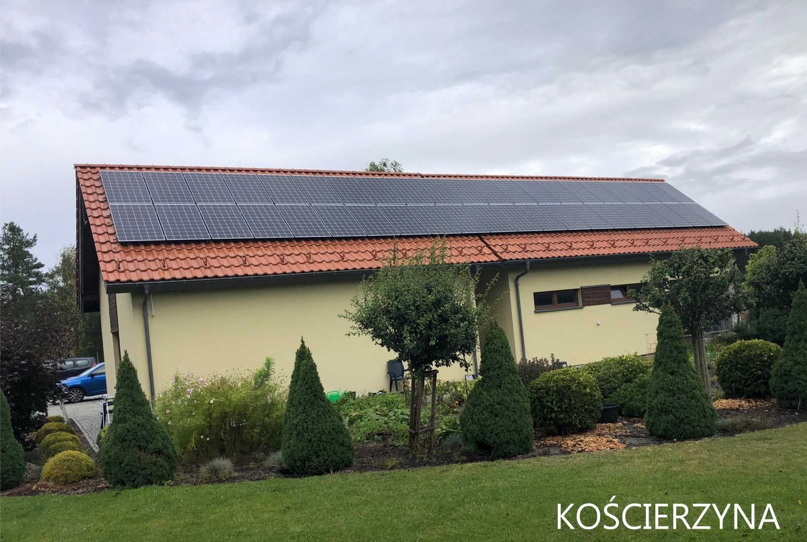 Dom jednorodzinny z panelami słonecznymi na czerwonym dachu, widok z poziomu ogrodu z ozdobnymi iglakami i krzewami, pochmurne niebo.