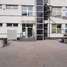 Pracownik w żółtym kasku na podnośniku koszowym, myjący okna na wysokości zewnętrznej windy w budynku biurowym