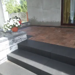 Schody wejściowe wyłożone czarną kostką, prowadzące do domu z brązową kostką brukową na tarasie, donica z białymi i czerwonymi kwiatami na murku obok schodów.