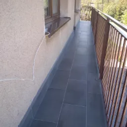 Długi balkon z szarymi, prostokątnymi płytkami podłogowymi, brązową balustradą i beżową elewacją; widoczny kabel elektryczny biegnący po ścianie.