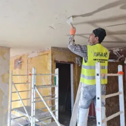 Pracownik w odblaskowej kamizelce, stojąc na rusztowaniu, szpachlą wyrównuje sufit w trakcie remontu. Widoczne niedoskonałości i ślady po instalacjach na ścianach.