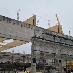 Budowa hali sportowej: surowy stan ścian z betonu komórkowego i żelbetu, widoczne zbrojenia, rusztowania i dźwig w tle, elementy konstrukcji drewnianej dachu.