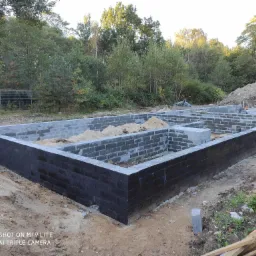 Wylany fundament z bloczków betonowych, zabezpieczony czarną izolacją, wypełniony piaskiem, widok z góry, drzewa w tle.