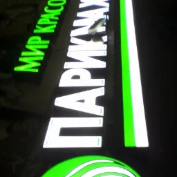 Podświetlany szyld reklamowy z zielonymi i białymi elementami: logo salonu piękności i nazwy w języku rosyjskim.