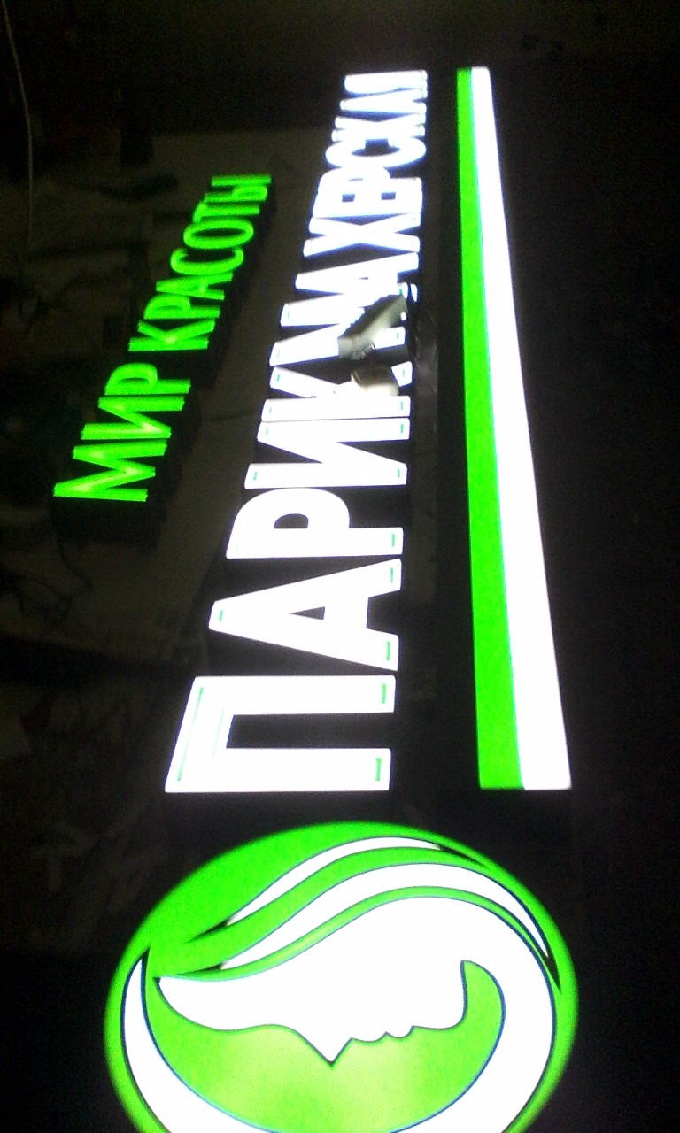 Podświetlany szyld reklamowy z zielonymi i białymi elementami: logo salonu piękności i nazwy w języku rosyjskim.