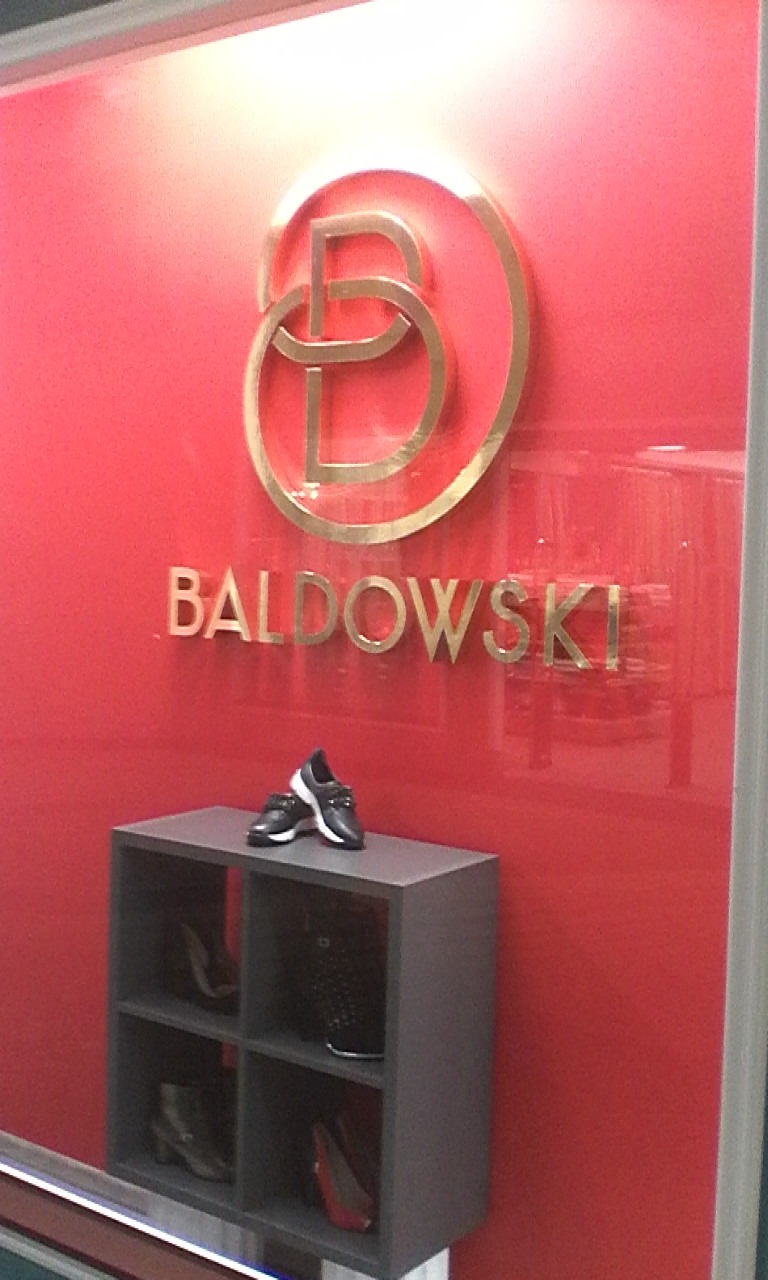 Złote logo firmy Baldowski na czerwonej ścianie, poniżej szara półka z butami, na górze półki para czarnych butów sportowych