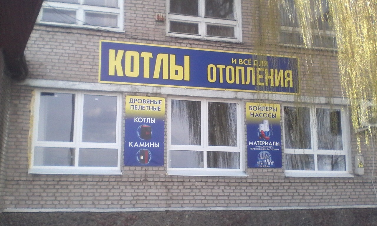 Budynek z cegły z szyldem reklamującym kotły i ogrzewanie, oraz banery w oknach z informacjami o kotłach, piecach i materiałach instalacyjnych.