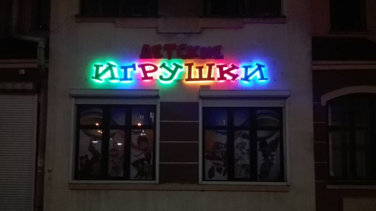 Fasada budynku z neonowym napisem 'ДЕТСКИЕ ИГРУШКИ' (Dziecięce Zabawki) nad dwoma oknami z grafikami postaci z bajek. Napis wykonany w różnych kolorach: zielonym, niebieskim, pomarańczowym...