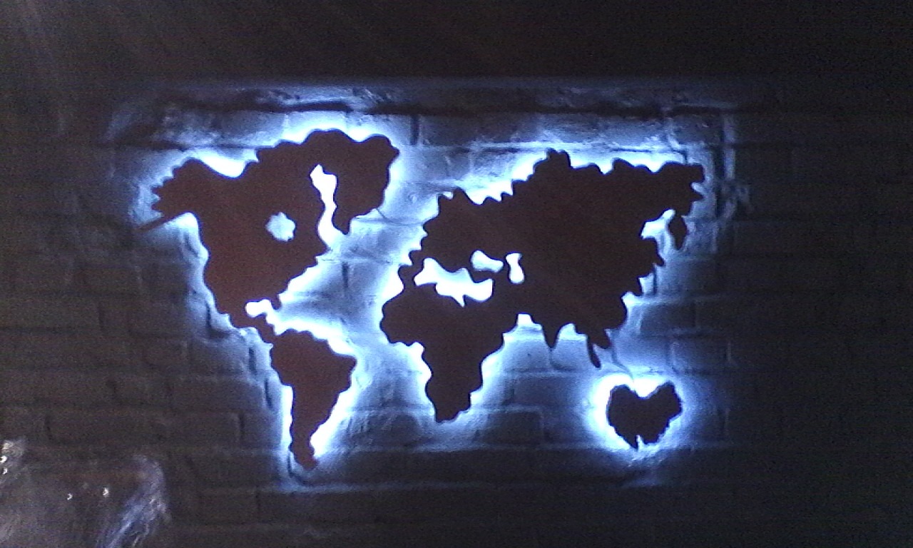 Świecąca mapa świata wycięta w ciemnej płycie zamontowanej na ceglanej ścianie, z niebieskim podświetleniem LED.