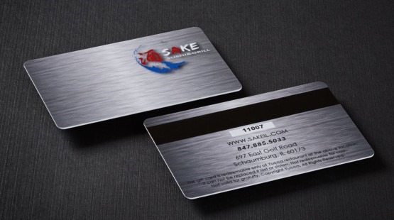 Dwie metalowe karty prezentowe z logo restauracji Sake Sushi & Grill, leżące na ciemnym tle, ukazują szczotkowane wykończenie i dane kontaktowe.