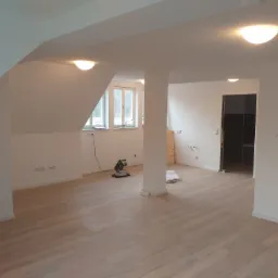 Jasne wnętrze poddasza z drewnianą podłogą w trakcie prac wykończeniowych, widoczna piła do cięcia paneli oraz kolumna nośna.