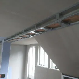 Montaż metalowej konstrukcji sufitu podwieszanego, widoczne profile i płyty gipsowo-kartonowe w surowym wnętrzu z oknami.