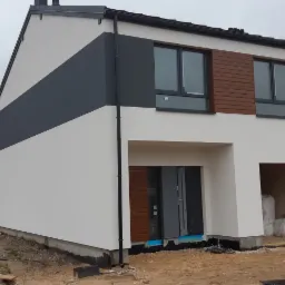 Nowoczesny szeregowiec z biało-szarą elewacją, drewnopodobnymi panelami i antracytowymi oknami, w trakcie wykończenia terenu.