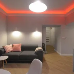 Jasne mieszkanie z szarą sofą, różowymi poduszkami, drewnianą podłogą i ozdobnym oświetleniem LED wokół sufitu.