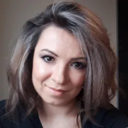 Portret uśmiechniętej kobiety z brązowymi włosami z pasemkami w czarnej bluzce, z ręką opartą na szarej dzianinie.