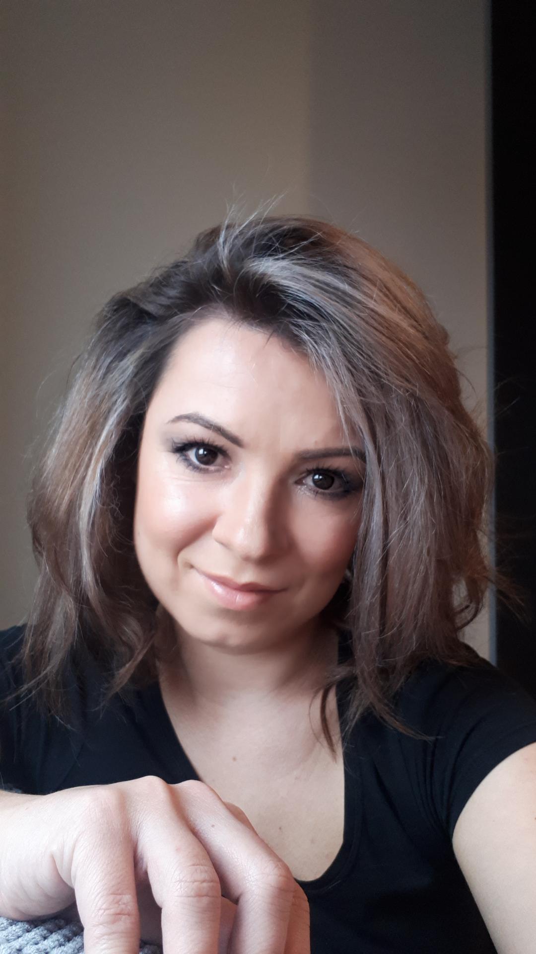 Portret uśmiechniętej kobiety z brązowymi włosami z pasemkami w czarnej bluzce, z ręką opartą na szarej dzianinie.