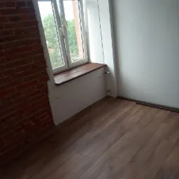 Wnętrze pokoju w trakcie prac wykończeniowych: ułożone panele podłogowe, fragment ściany z cegły, okno z drewnianym parapetem, biała ściana.