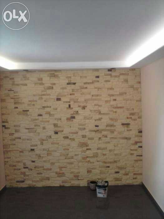 Ściana wyłożona dekoracyjną okładziną imitującą cegłę, z widocznym oświetleniem LED podsufitowym i wiaderkiem z narzędziami w rogu.